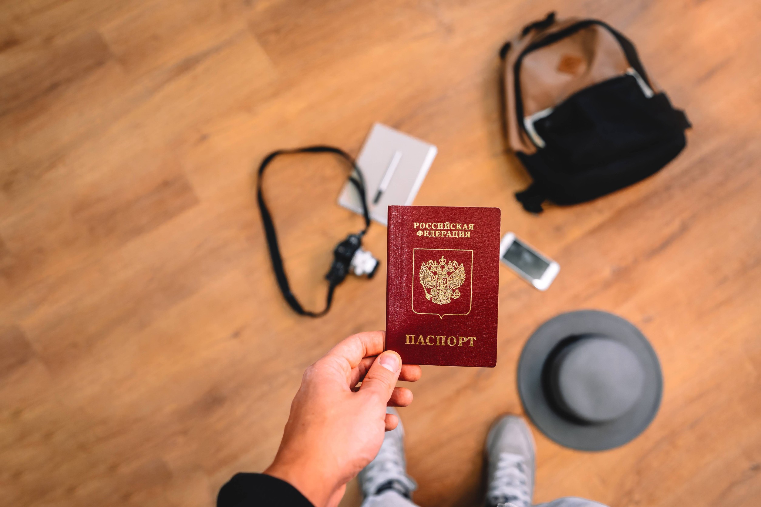 Man Holds A Russian Passport With A Backpack And Travel Accessories In The Background